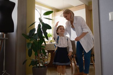 Gülümseyen genç bir anne kızının evde okula hazırlanmasına yardım eder. Anne cesaret verirken çocuk okul üniforması giydiği için heyecanlı görünüyor..