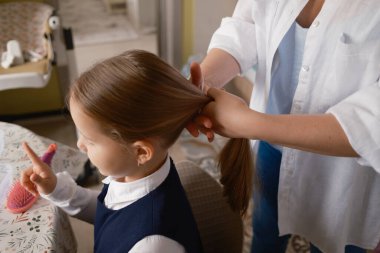 Bir anne, sabah okulu rutininin bir parçası olarak kızının saçını ustalıkla örer. Çocuk sabırla oturur, bir şeye işaret eder, hassas, günlük aile anlarını vurgular..