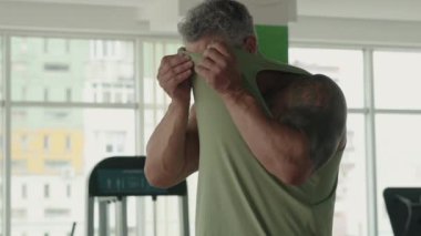 Adam spor salonunda egzersiz yaparken terini silmek için tişörtünü kullanıyor. Fitness ve sıkı çalışmaya olan bağlılığını gösterir.