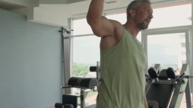 Yeşil kollu tişörtlü kaslı adam, güç antrenmanına başlamadan önce modern bir spor salonunda ısınma egzersizleri yapıyor..