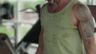 Spor salonunda dambıllarla çalışan, yeşil kolsuz tişörtlü, fitness ve güç antrenmanlarına odaklanmış kaslı bir adam..