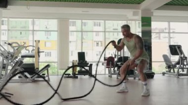 Bir adam, modern bir spor salonunda savaş halatlarıyla yoğun bir idman yapar. Güç ve zindelik üzerine yoğunlaşır..