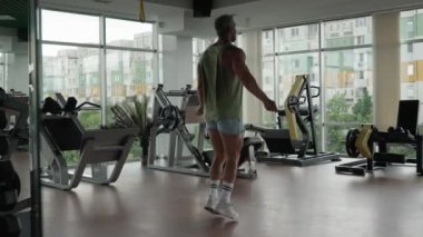 Atletik adam geniş, iyi donanımlı bir spor salonunda ip atlıyor. Şehir manzarasına bakan geniş pencereleri var..