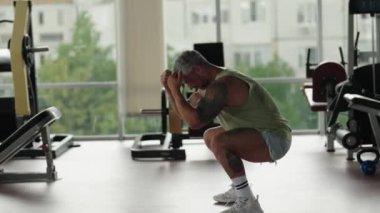 Dövmeli atletik adam, büyük pencereli ve modern spor aletli iyi donanımlı bir spor salonunda çömelme egzersizi yapıyor..