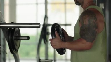 Kolu dövmeli bir adamın yakın plan çekimi. Spor salonundaki halterin üzerine fazladan ağırlık koyuyor. Güç ve zindeliği vurguluyor..