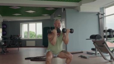 Dambıllarla çömelmiş bir adam, iyi donanımlı bir spor salonunda güç ve spor eğitimi sergiliyor. Kas geliştirmek ve fiziksel sağlığı geliştirmek için egzersiz..