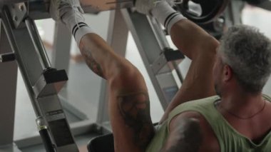 Dövmeli bir adam spor salonunda bacak pres makinesinde çalışıyor, bacak dayanıklılığına ve forma odaklanıyor..
