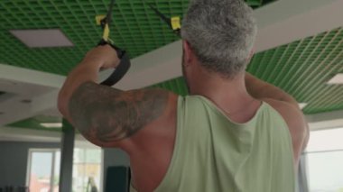 Olgun bir adam spor salonunda süspansiyon kayışlarıyla çalışıyor, güç ve kas gelişimine odaklanıyor. İçerde egzersiz yapan daha yaşlı bir yetişkinin arka görüntüsü..