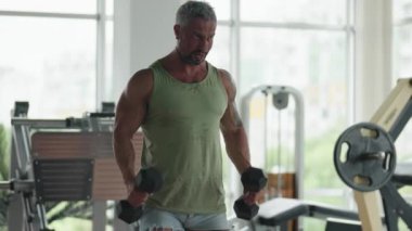 Spor salonunda spor yapmak için dambılları kaldıran bir adam. Güç antrenmanı ve kas geliştirmeye odaklanmış durumda..