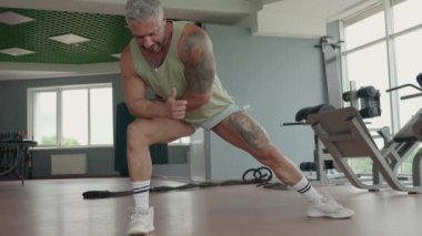 Spor salonunda kaslı bir adam antrenmandan önce ısınma egzersizi yapıyor. Esniyor, antrenmana hazırlanıyor ve kaslarının hazır olduğundan emin oluyor..