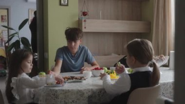 Erkek ve kız kardeşler okuldan önce mutfak masasında sağlıklı bir kahvaltı yapıyorlar. Okul günü için hazırlanırken taze meyve yiyorlar..