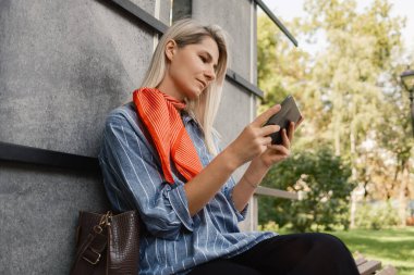 Açık havada oturan kırmızı eşarplı, rahat bir günde akıllı telefonuyla meşgul olan rahat ve modern bir park ortamındaki bağlantının göstergesi olan sıradan giyinmiş bir kadın..