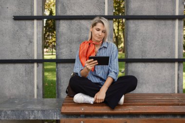 Tabletine sarılmış bir kadın bankta bağdaş kurmuş oturuyor. Arka plan, modern estetiğe sahip huzurlu bir park ortamı olduğunu gösteriyor..