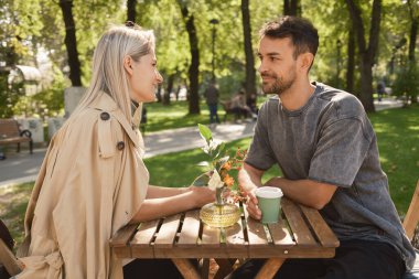 Bir çift, güneşli bir günde parktaki ahşap bir masada kahve içip sohbet ediyorlar. Ayar rahat ve yeşillikle dolu, bir bağlantı anı yakalıyor..