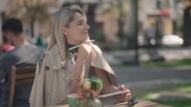 Sarışın bir kadın kafeterya masasında oturur ve güzel bir günde dışarıda stili olan bir tablet kullanır. Gülümsüyor, mutlu ve düşünceli..