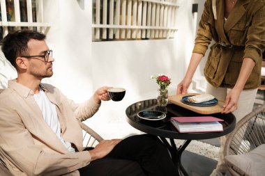 Bir adam elinde kahve fincanıyla açık bir kafe masasında otururken garson tepside yemek getiriyor. Rahatlatıcı ortam ya sakin bir yemek ya da ara vermeyi öneriyor..