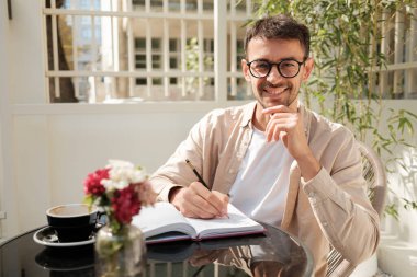 Neşeli bir adam açık bir kafe masasında oturur, bir deftere yazar ve kahvenin tadını çıkarır. Yeşilliklerle çevrili, rahat ve odaklanmış görünüyor, bir ilham anı yakalıyor..