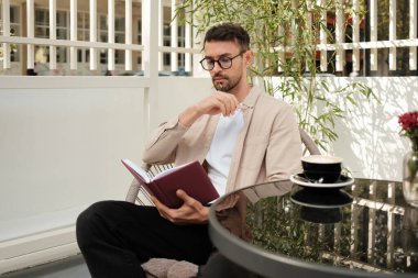 Gözlüklü düşünceli bir adam açık bir kafede bir fincan kahveyle çevrili bir şekilde kitap okuyor..