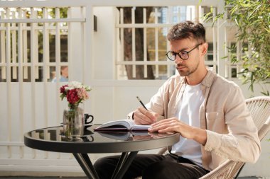 Açık bir kafe masasında otururken not defterine yazan düşünceli bir adam. Huzurlu ve huzurlu bir manzara, çiçekler doğaya bir dokunuş ekliyor..