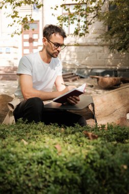 Beyaz tişörtlü ve gözlüklü bir adam bir kütüğün üzerine oturur ve bir deftere yazar. Düşünceli ve meşgul görünüyor. Etrafı yeşillik ve sıcak güneş ışığıyla çevrili..
