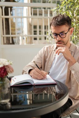 Gözlüklü bir adam açık bir kafede düşünceli bir şekilde oturur ve bir deftere yazar. Ayar parlak ve rahat. Masada çiçekler var. Rahatlamış bir atmosfer var..