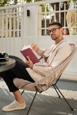 Genç bir adam, kendini kitaplara kaptırmış, modern bir açık hava sandalyesinde rahatça oturuyor. Çevresindeki ağaçların arasından süzülen doğal ışıkla huzurlu bir ortam yaratmayı seviyor..