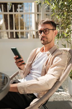 Gözlüklü rahat bir adam akıllı telefonunu kullanarak teras sandalyesine oturur. Güneşli açık hava manzarası, sakin ve sakin bir atmosfer yaratan yeşillik ve modern mimariye sahiptir..