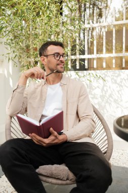 Düşünceli bir adam, gündelik giysiler içinde bir bahçede oturmuş, elinde bir kitap ve kalemle, düşüncelerini yansıtıyor. Güneşli açık hava, sakin ve düşünceli bir atmosfer ekler..
