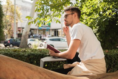 Gözlüklü genç bir adam güneşli bir günde dışarıda oturur, elinde bir tablet tutarken düşünür. Arka planda arabalar ve yeşillik olan kentsel çevre, rahatlama ve yansımayı öneriyor.
