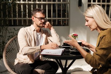 İki profesyonel açık hava kafeteryasında gündelik bir tartışma içindeler. Not defterleri ve kahveyle, şık ve güneşli bir ortamda ortak bir toplantıdan hoşlanıyorlar.