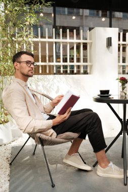 Genç bir adam teras sandalyesinde oturur, kahve içerken kitap okur. Ortalık sakin ve rahatlatıcı. Doğal ışık ve yeşillik onu çevrelemiş..