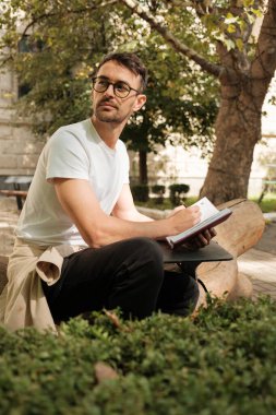 Genç bir adam düşünceli bir şekilde bir bankta oturur ve defterine doğayla iç içe bir şeyler yazar. Ayarlama yaratıcılığı, rahatlığı ve dışarıyla bağlantıyı iletir..