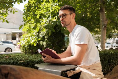 Düşünceli bir adam dışarıda oturur, yemyeşil ağaçlarla çevrili bir deftere yazı yazar. Sıcak güneş ışığı, yaratıcılık ve yansıma için ideal sakin ve düşünceli bir atmosfer yaratır..