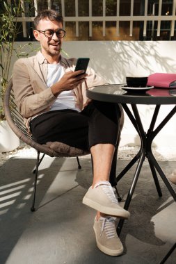 Neşeli bir adam açık bir kafede oturmuş, akıllı telefonunun keyfini çıkarıyor. Güneş ışığı sıcak ve rahat bir atmosfer yaratır, rahatlık ve iletişim için mükemmeldir..