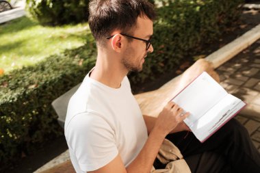 Beyaz gömlekli genç bir adam güneşli bir günde yemyeşil bir bahçede bir kitap okuyor. Rahatlamış ve odaklanmış görünüyor..