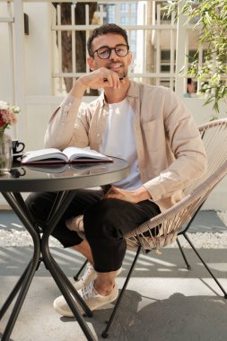 Açık bir kafe masasında oturmuş kitap okuyor ve huzurlu bir anın tadını çıkarıyor. Ortam rahat ve güneşli, huzur ve rahatlık hissi katıyor..