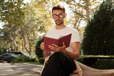 Gözlüklü bir adam dışarıda, huzurlu bir park ortamında oturmuş güneşli bir günün dinginliğinin ve temiz havasının tadını çıkarırken kitap okuyor..
