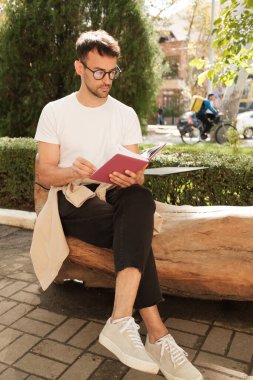 Dışarıda bankta oturan genç bir adam, doğal ışık altında kitap okumaya odaklanıyor. Görüntü bir rahatlama, konsantrasyon hissi veriyor ve huzurlu bir anın tadını çıkarıyor..