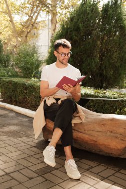 Gözlüklü genç bir adam parkta bir bankta oturup kitap okuyor. Gün ışığı ağaçların arasından akarken ortam huzurlu..