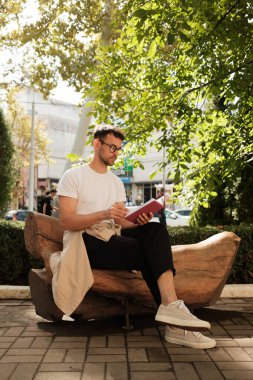 Bir adam parkta ahşap bir bankta oturup kitap okurken güneşin tadını çıkarıyor. Yaprakların arasından süzülen güneş ışığıyla ortam huzurlu ve rahatlatıcı..