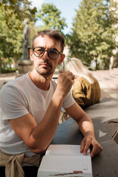 A thoughtful man in glasses holds a pen at his lips, pondering ideas in an outdoor park. Sunlight filters through the trees, creating a serene and contemplative atmosphere.
