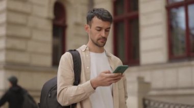 Sırt çantalı genç bir adam binanın önünde telefonunu kontrol ediyor. Bu video seyahat, iş ve günlük hayatın temalarını çağrıştırıyor.