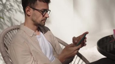 Genç bir işadamı sandalyede oturuyor ve telefonunu kullanıyor. Telefonuna bakarken gülümsüyor. Sıradan giyinmiş..