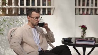 Kafe masasında hasır bir sandalyede oturan bir adam kahve içiyor. Bir garson ona bir dilim cheesecake getiriyor. O, gözlük, açık kahverengi bir gömlek ve koyu pantolon giyiyor..
