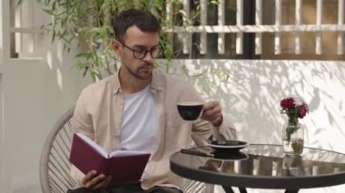 Adam kitap okuyor ve açık bir kafede kahve içiyor. Kitabını okurken bir yudum kahve içer..