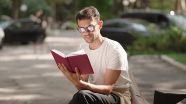 Genç bir adam parkta bir bankta oturur ve kitap okur. Beyaz bir tişört ve gözlük takıyor. Arkadan arabalar geçiyor..