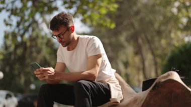 Genç bir adam parkta bir kütüğe oturmuş, telefonunu kullanıyor. Beyaz bir tişört ve gözlük takıyor ve kısa kahverengi saçları var. Arkaplan bulanık.