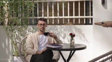 Bir adam açık bir kafede bir masada oturuyor, kahvesini yudumluyor. Sunucu ona bir tabak suşi getiriyor. Güneş ışığı bitkilerin arasından akar ve rahatlatıcı bir atmosfer yaratır..