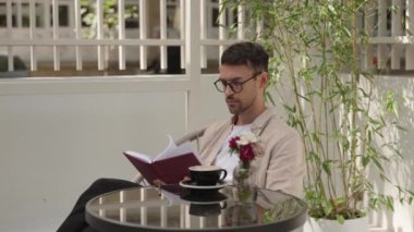 Adam kitap okuyor ve açık kafede kahve içiyor. Güneşli bir günde okumayı ve kahve içmeyi seviyor..