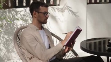 Açık kafede deftere yazan bir adam. O, gözlük ve bej bir gömlek giyiyor. Güneş ışığı ve bitkilerin gölgeleri görülebilir..